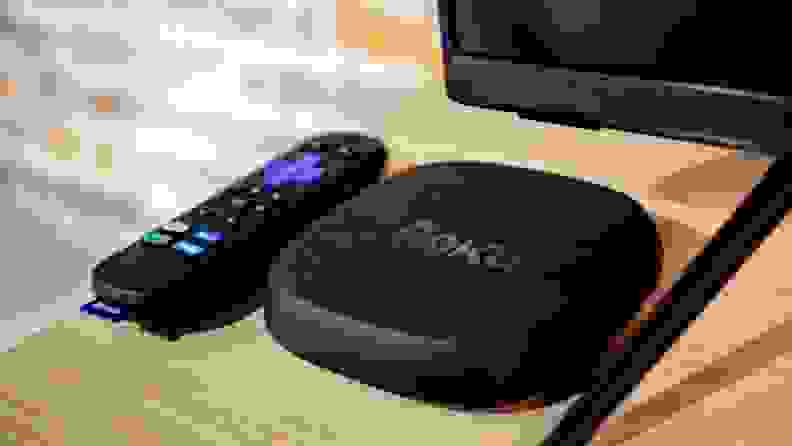 The black and rectangular Roku Ultra sits next to its black and multi-buttoned remote beneath the corner of a TV.