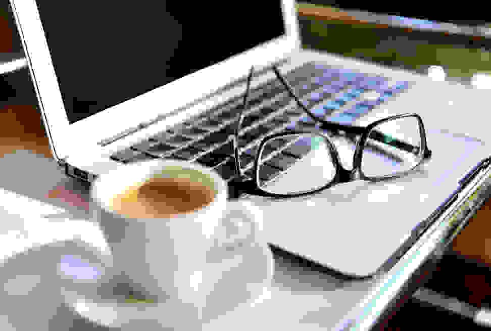 A laptop, hot beverage, and eyeglasses on a desk.