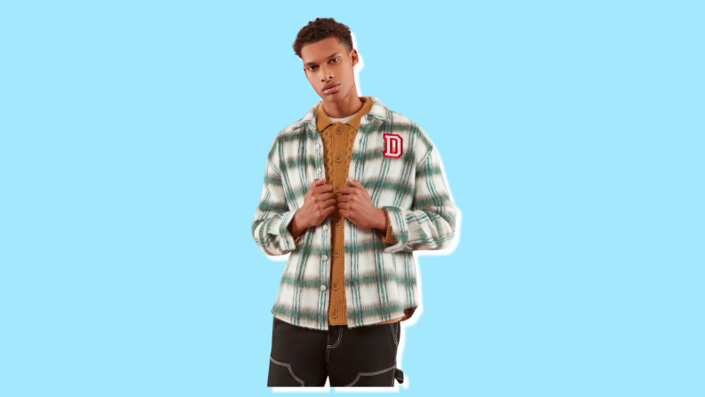 A man stands facing the camera, wearing a green and white plaid flannel shirt with an embroidered "D" on the left pocket area.