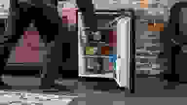一名男子走进镜头，进入一个开放的Insignia NS-CF26BK9迷你冰箱内部。里面装满了食物和饮料。