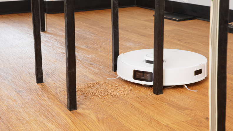 Small white robot vacuum on top of hardwood floors under skinny constructed table legs in front of pile of cork shavings.