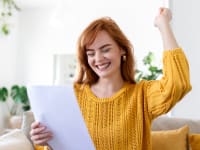A woman in a yellow sweater smiles and raises her fist while reading a document, celebrating good news.