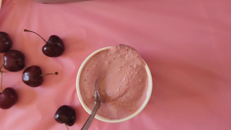 Cherry ice cream inside of a small bowl with a spoon inside next to several cherry fruits.