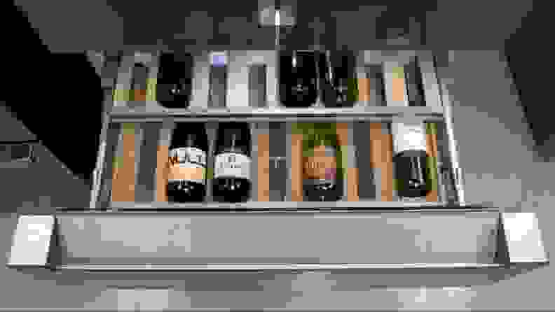 A close-up of the open refreshment center drawer.The glass-bottom drawer is lined with wooden slats and both the lower and upper shelves are holding several wine bottles.