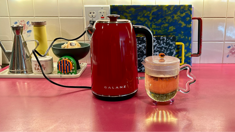 A Galanz electric kettle in the color red, next to a small, clear, glass, teapot.