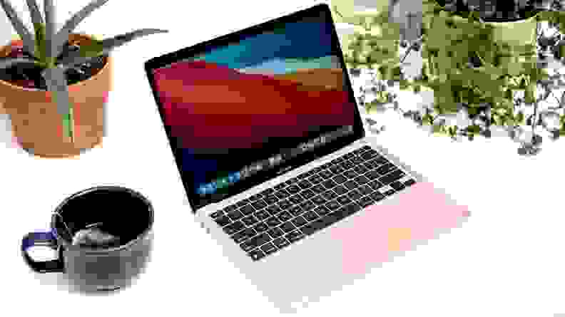 A rose gold colored laptop on a white desk surrounded by plants and a coffee cup.