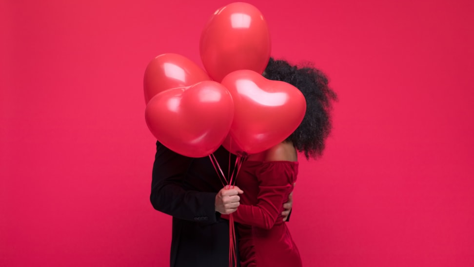 A sharply dressed couple kissing, faces obscured by heart balloons; against a red background
