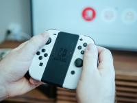 Close-up of the Nintendo Switch OLED controller with white Joy-Con attached on either side. The Switch menu UI is displayed on the TV in the background.