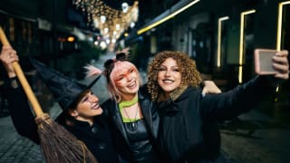 Three young women in witch costumes taking selfies on a city street during nighttime