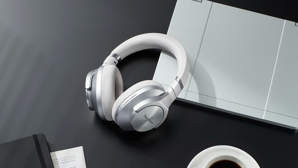 A pair of shiny, silver and white headphones sit on a stylish black desk next to computing peripherals.