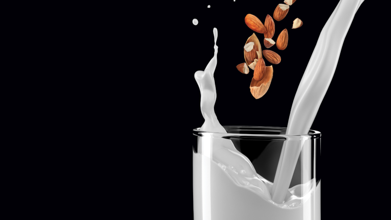 A person is pouring almond milk into a glass, making a splash of milk and almond pieces.
