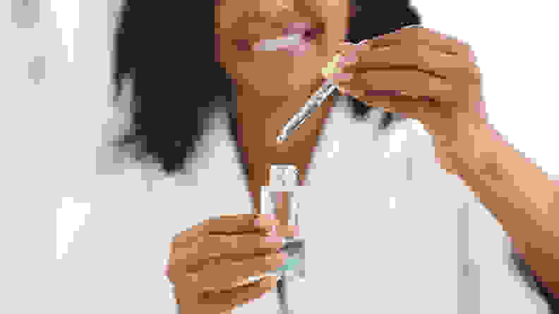 A woman holds a bottle of blue liquid in one hand and a dropper in the other hand and smiles.