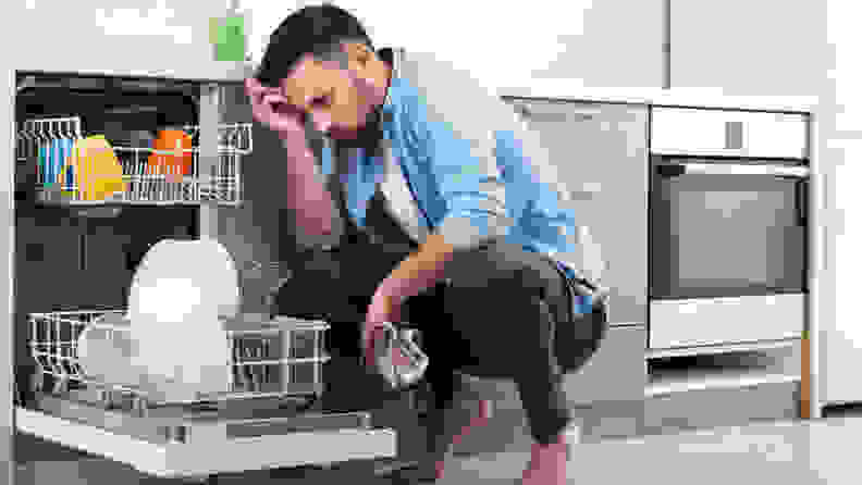 A man crouches in front of an open dishwasher, holding a dish towel, looking visibly upset.