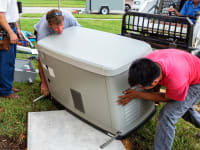 A group of men are moving a standby generator off of a fork lift and onto a platform set up in someone's yard.