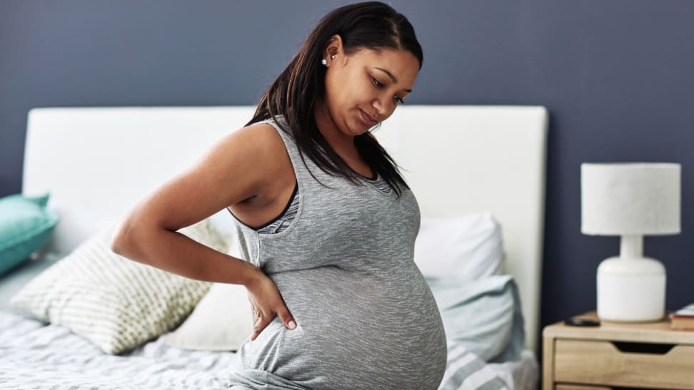 Pregnant person sitting on bed with hand on back in discomfort.