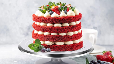 A four-layered strawberry cake, red with white icing, is displayed on an aluminum cake stand.