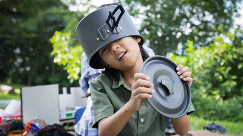 Kid with a outdoor cooking pan on her head is holding the lid of the pan in a park.
