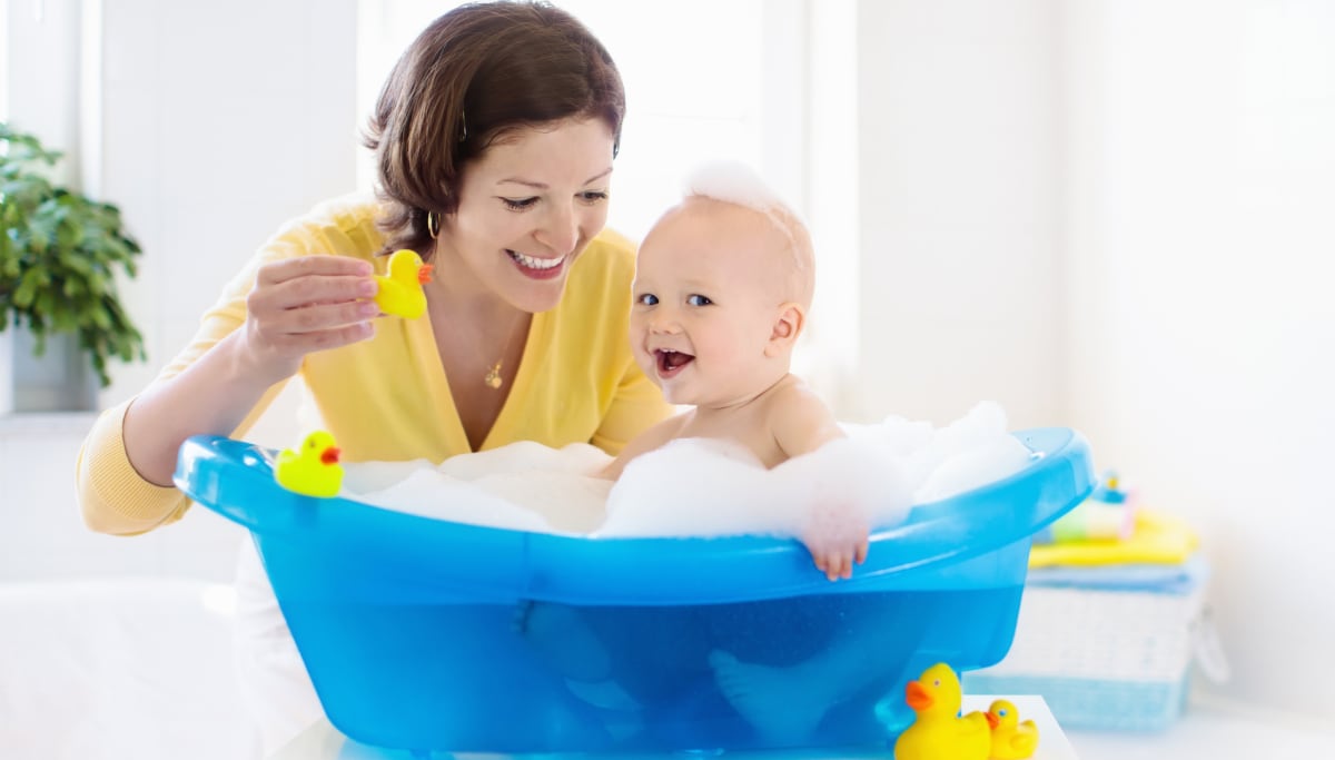 madre bañando a su bebé en una bañera azul, con patitos de goma y agua con mucha espuma