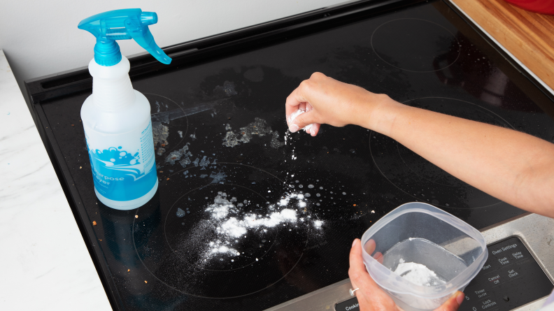 A hand sprinkling baking soda on a glass stove.