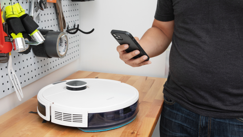 Person hovering smartphone closely to white circular robot vacuum sitting on wooden workbench in front of pegboard with tools.