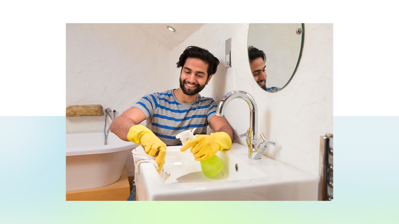 A person in yellow cleaning gloves happily wipes down a bathroom sink with a towel and spray bottle in hand.