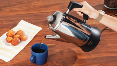 A person's hand pouring coffee from an electric coffee percolator into a blue mug