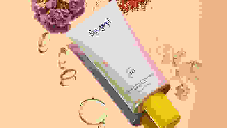 A white tube of sunscreen on a peach background with ingredients laying around it.