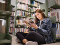 Student sitting on floor of the library, leaning against bookshelf and reading  a textbook