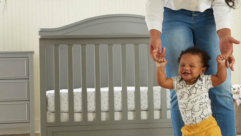 A smiling toddler walks in front of their convertible baby crib