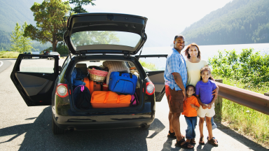 A family of four stands next to their car loaded up with suitcases.