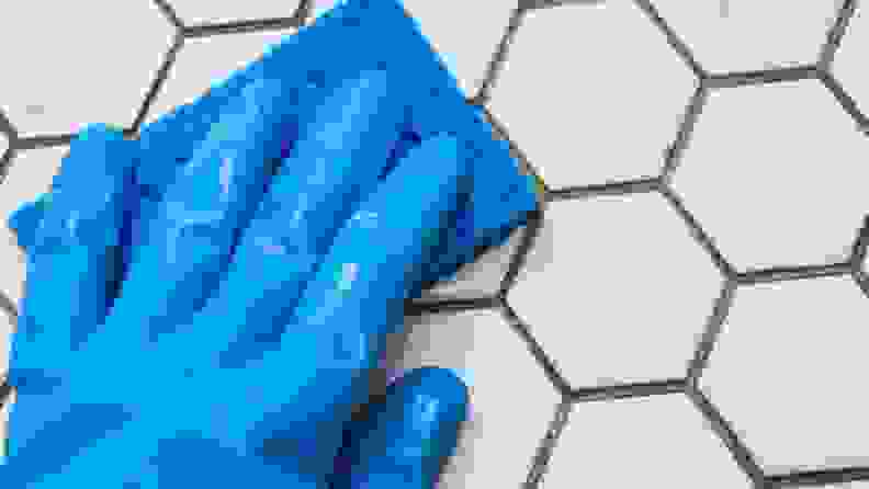 Hand wearing blue rubber gloves and blue sponge to wipe tile surface.