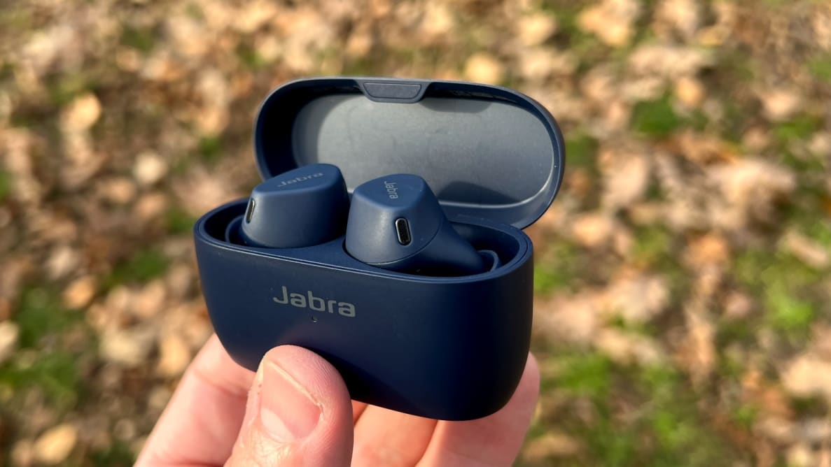 A pair navy earbuds sit in their open charging case, held in a hand before a leaf-covered ground.