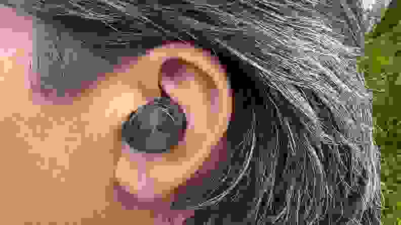 The gunsmoke colored wireless earbud sits in an ear of a person with salt and pepper brown hair.