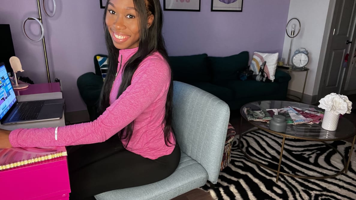 Person smiling while sitting on top of blue office chair with wide seat cushion in front of pink desk while one hand rests on laptop trackpad inside of colorful living room.