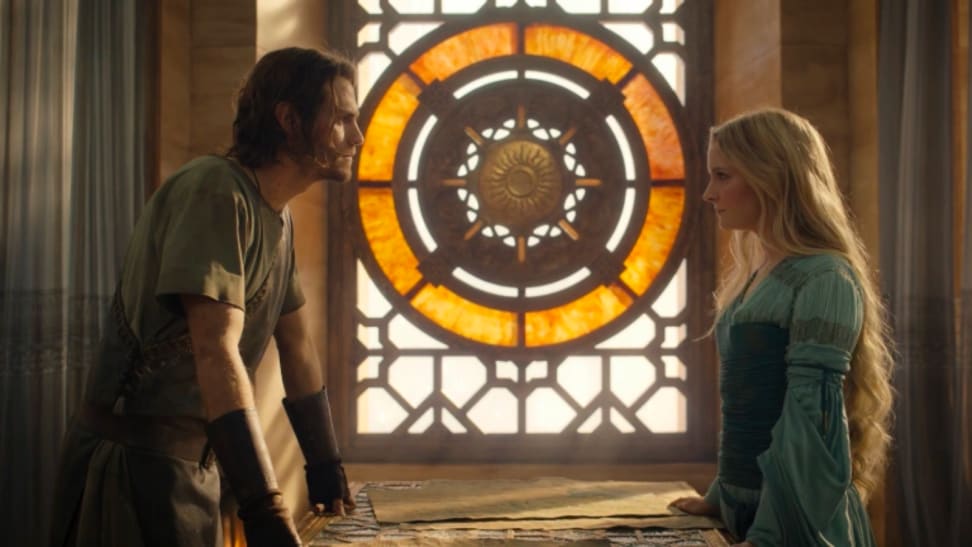 A still from 'The Rings of Power' that features Halbrand and Galadriel standing across fromo one another at a table, before a stained glass window.