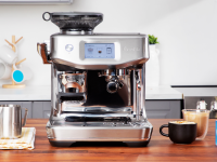Breville Barista Touch Impress espresso machine in stainless steel finish sitting on top of wooden countertop next to two cups of coffee, an acrylic paperweight and a steel frothing pitching.
