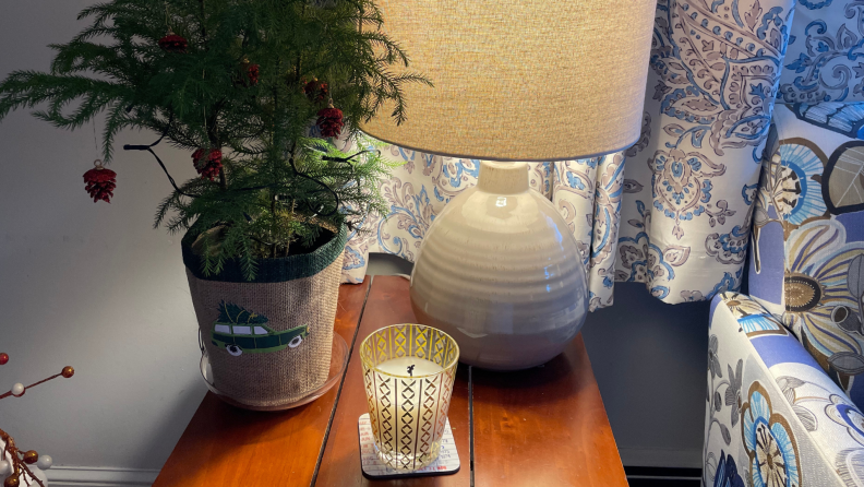 A candle on a side table with a lamp and mini tree.