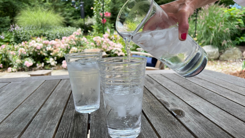 A hand pouring ice water from a pitcher into a glass on an outdoor table with another glass in the background.