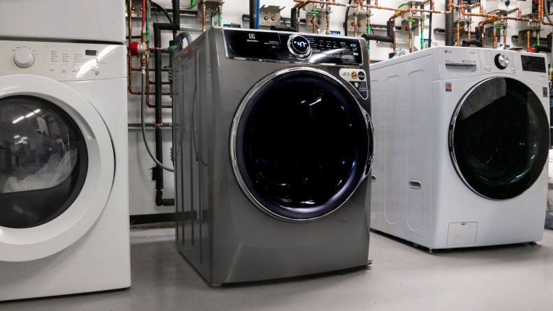 A close-up of the Electrolux ELFW7637AT front-load washing machine between two other washers in our laundry testing lab. Behind the washers you can see a network of pipes supplying each unit with temperature-regulated water.