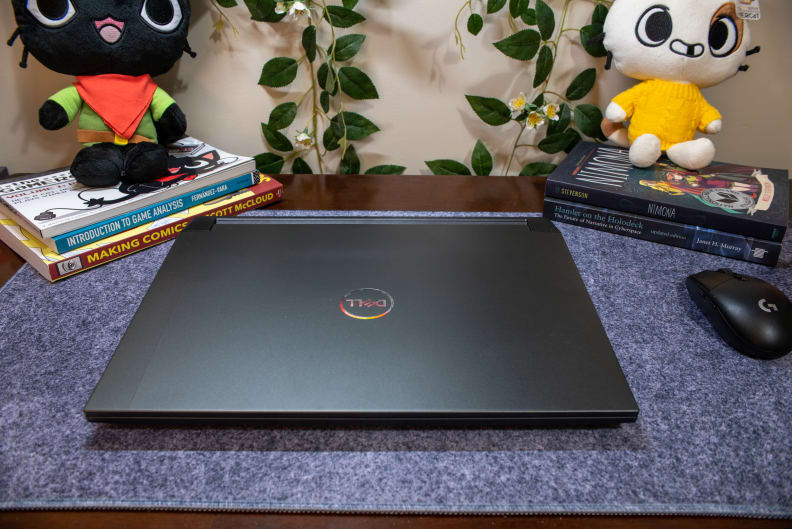 A black laptop, close, on top of a blue-gray mat flanked by books and plushies.