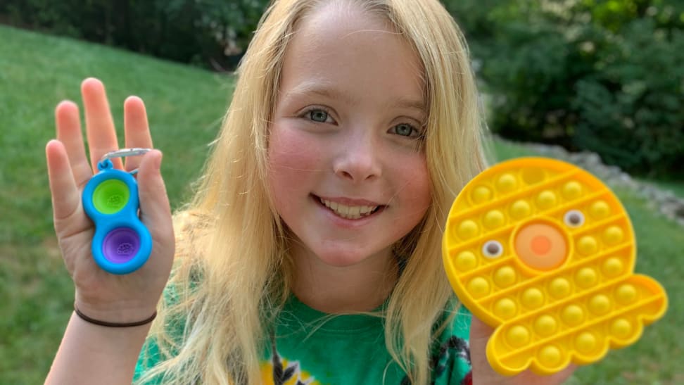 a little girl hold's up two pop-it fidget toys, one small 2-button one and another larger squid-shaped one