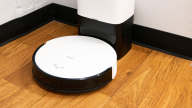 A round white robot vacuum sits in its white dock in the corner of a white room with brown wood floors