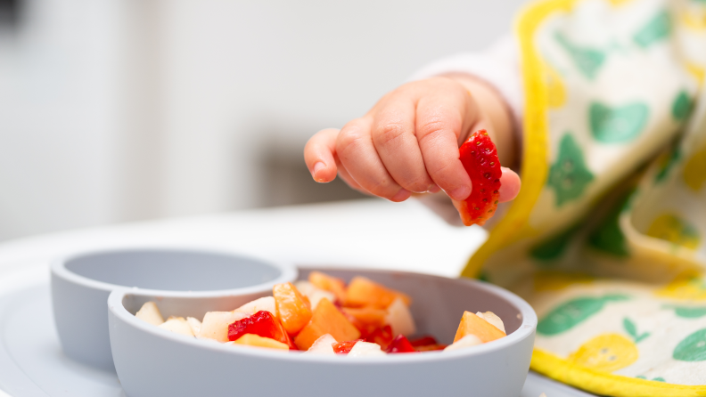 Bowl of cut up strawberries with a baby's hand holding a piece of one.