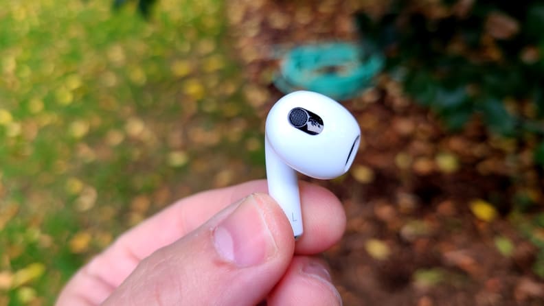 The all-white AirPod sits in the reviewer's hand with its side vent facing the camera before grass and wood chips in the background.