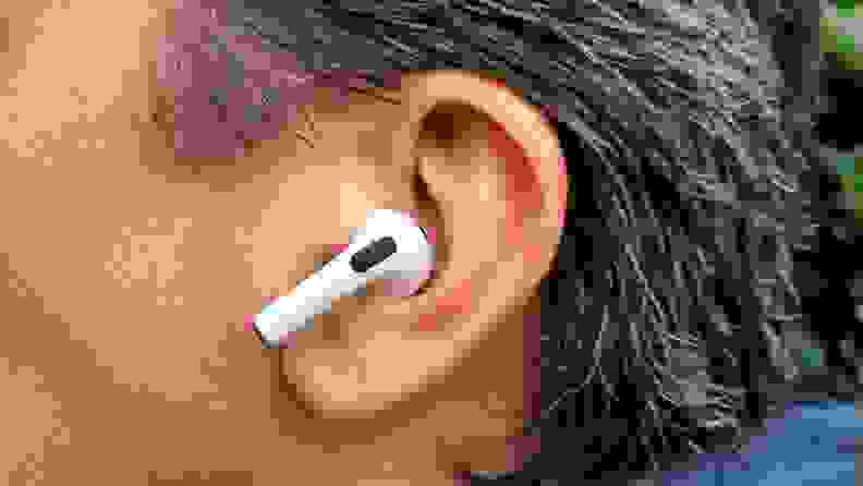 The all-white AirPod sits in the reviewer's ear with its shortened stem facing forward, set above a gret sweatshirt hood and below brown grey hair.