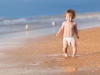 A baby in a diaper walking in the sand.