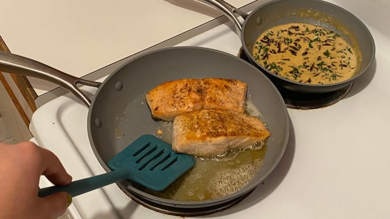 A person uses a spatula to flip salmon fillets in a nonstick skillet.