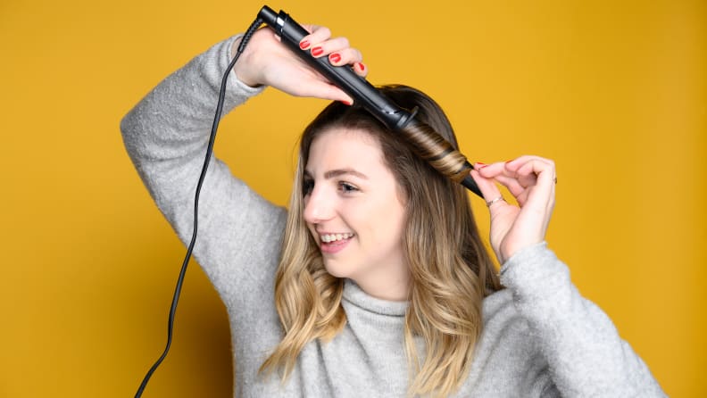 A person with long hair holds a curling iron in their hair and smiles.