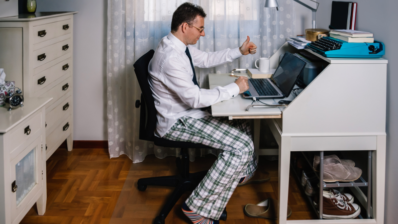 While working from home, a laborer wears a dress shirt and tie as well as pajama pants and slippers. Over a video meeting on their laptop, they give a thumbs-up.