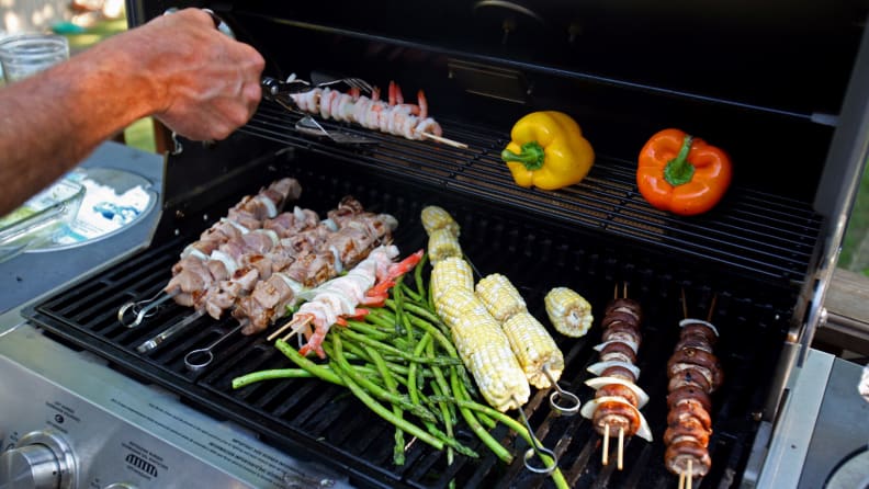 A hand adjusting food on a crowded gas grill, filled with meats and veggies